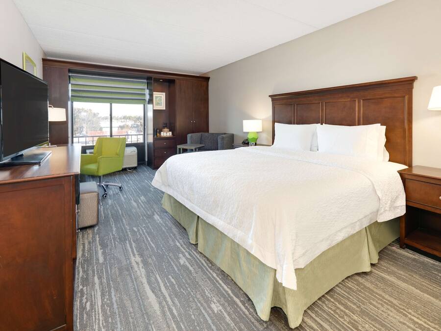 A hotel room with a large bed, a desk with a TV, a chair, and a window with blinds.