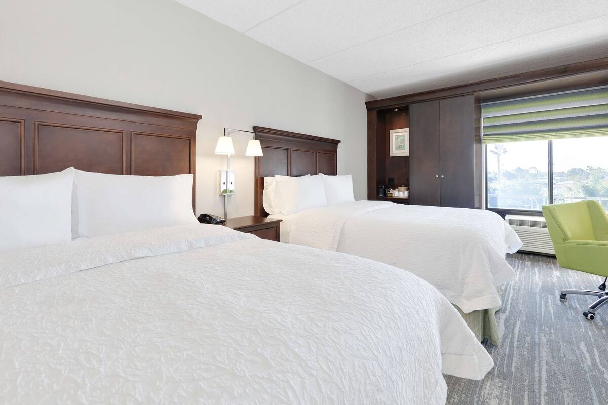 A hotel room with two beds, a nightstand with a lamp, a green chair, and a large window with blinds.