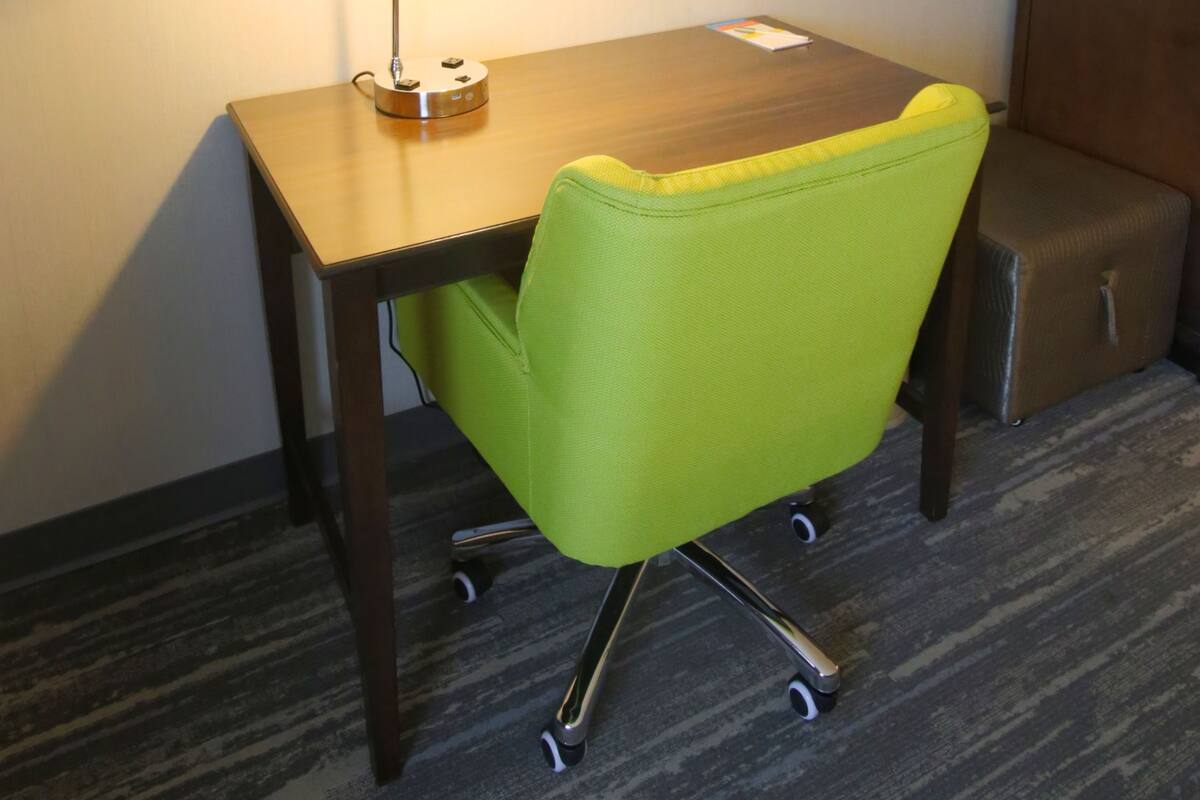 A hotel room with a desk, a green chair, a lamp, a framed picture on the wall, and a small cabinet.
