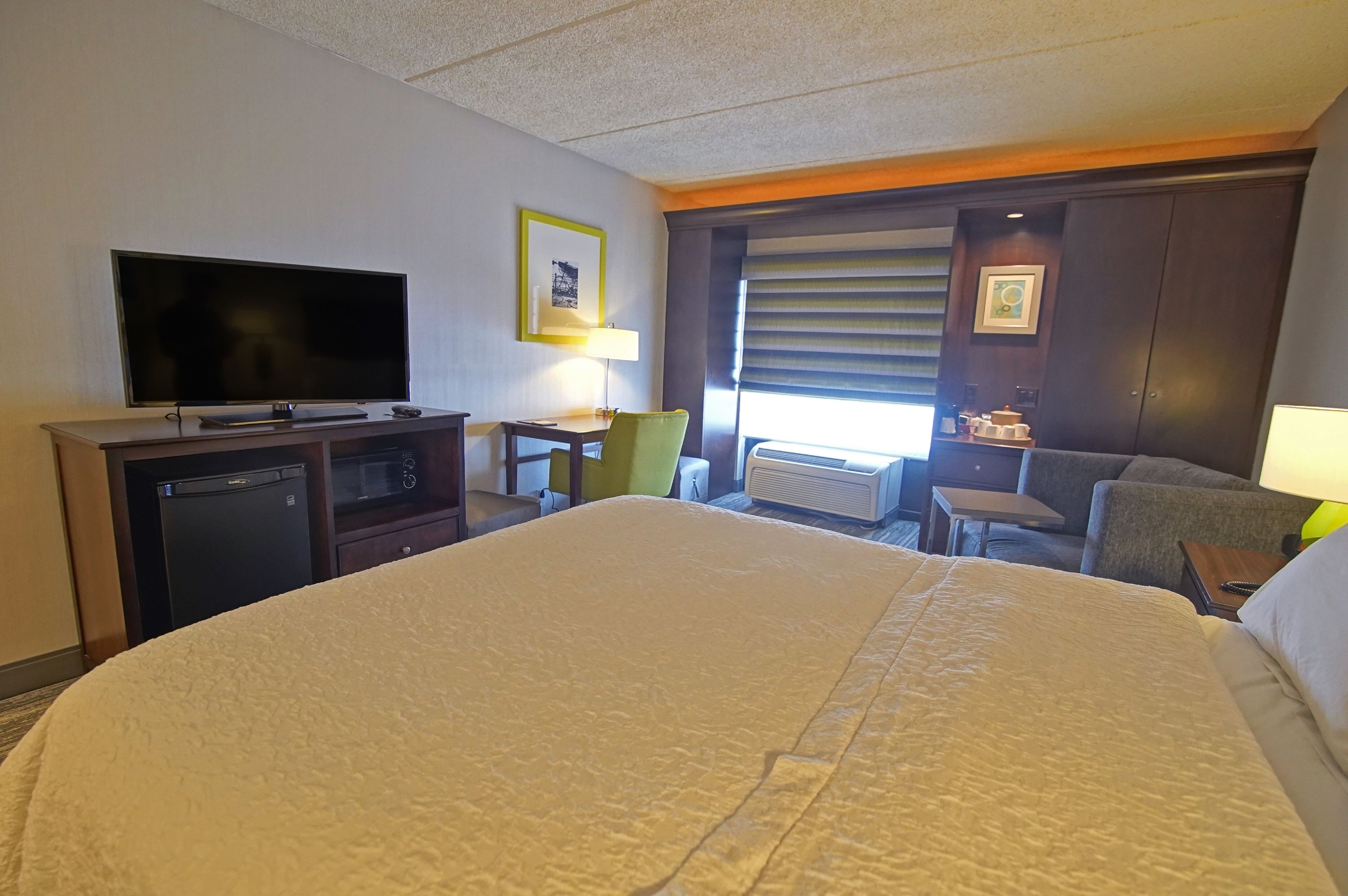 Una habitación de hotel con una cama grande, televisión de pantalla plana, escritorio con lámpara, una silla, una mesita y ventana con persianas.