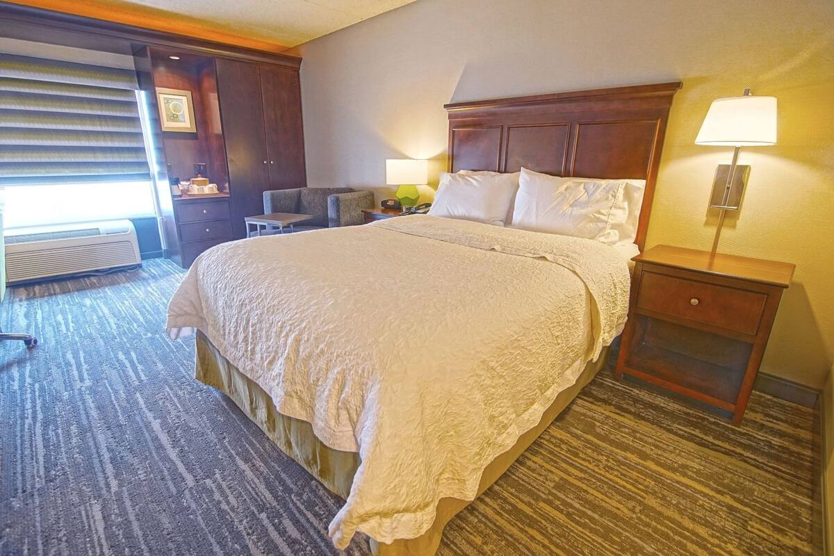 A hotel room with a large bed, a nightstand, a lamp, a desk, and a chair.
