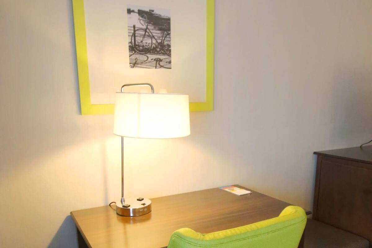 A hotel room with a desk, a green chair, a lamp, a framed picture on the wall, and a small cabinet.