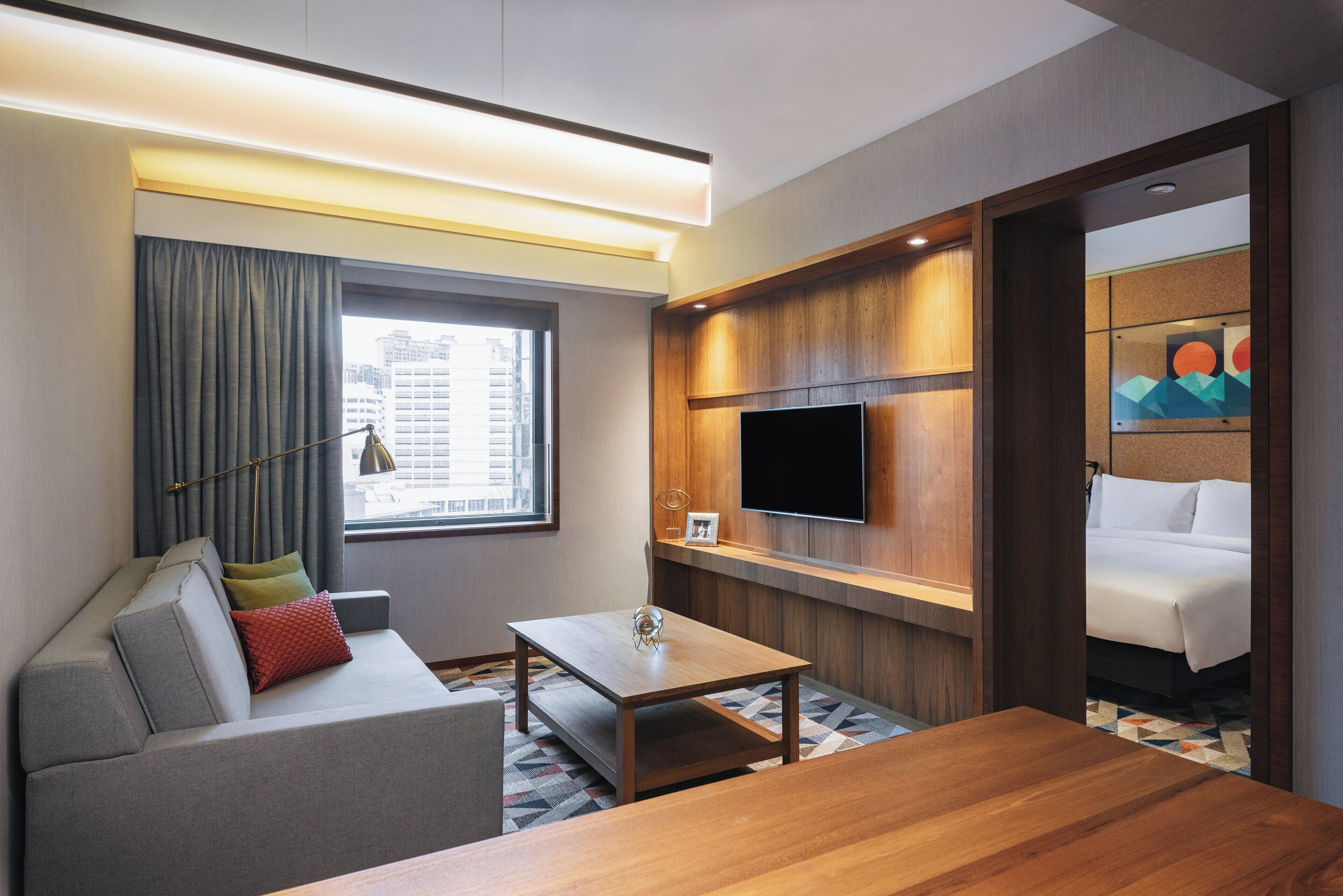 Quarto de hotel moderno com sofá, mesa de centro de madeira, TV de tela plana fixada em um painel de madeira e uma cama com edredom branco.