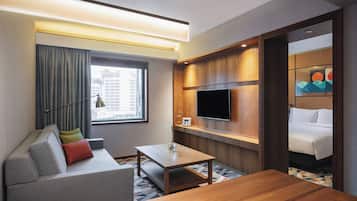 Quarto de hotel moderno com sofá, mesa de centro de madeira, TV de tela plana fixada em um painel de madeira e uma cama com edredom branco.