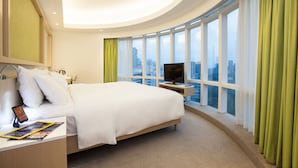 A modern hotel room with a large bed, a TV on a stand, a bedside table with a phone and a magazine, and a view of the city through large windows. - Eaton HK (Kowloon)