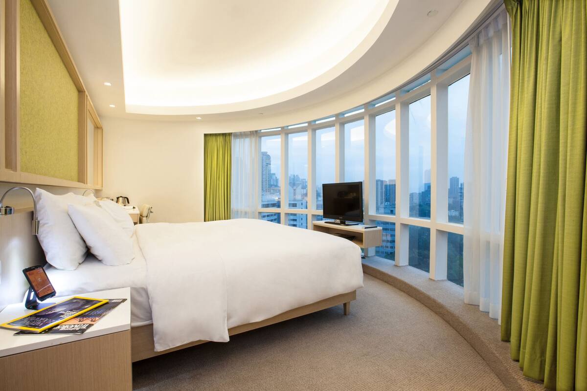 A modern hotel room with a large bed, a TV on a stand, a bedside table with a phone and a magazine, and a view of the city through large windows.