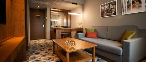 Una habitación de hotel moderna con una mesa de centro de madera, un sofá gris con cojines coloridos, una alfombra estampada y un espejo grande sobre el sofá.