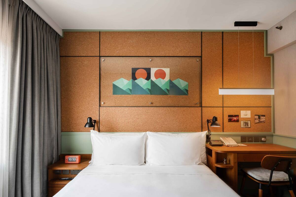 A hotel room with a bed, a desk with a chair, a corkboard wall with artwork, and a nightstand with a lamp.