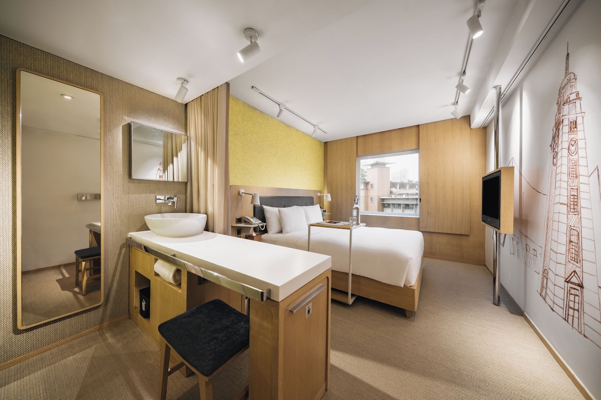 A hotel room with a large mirror, a sink, a bed, a bedside table, and a TV.