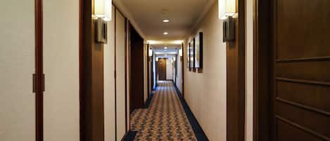 Pasillo de hotel con alfombra estampada, puertas de madera e iluminación en la pared.