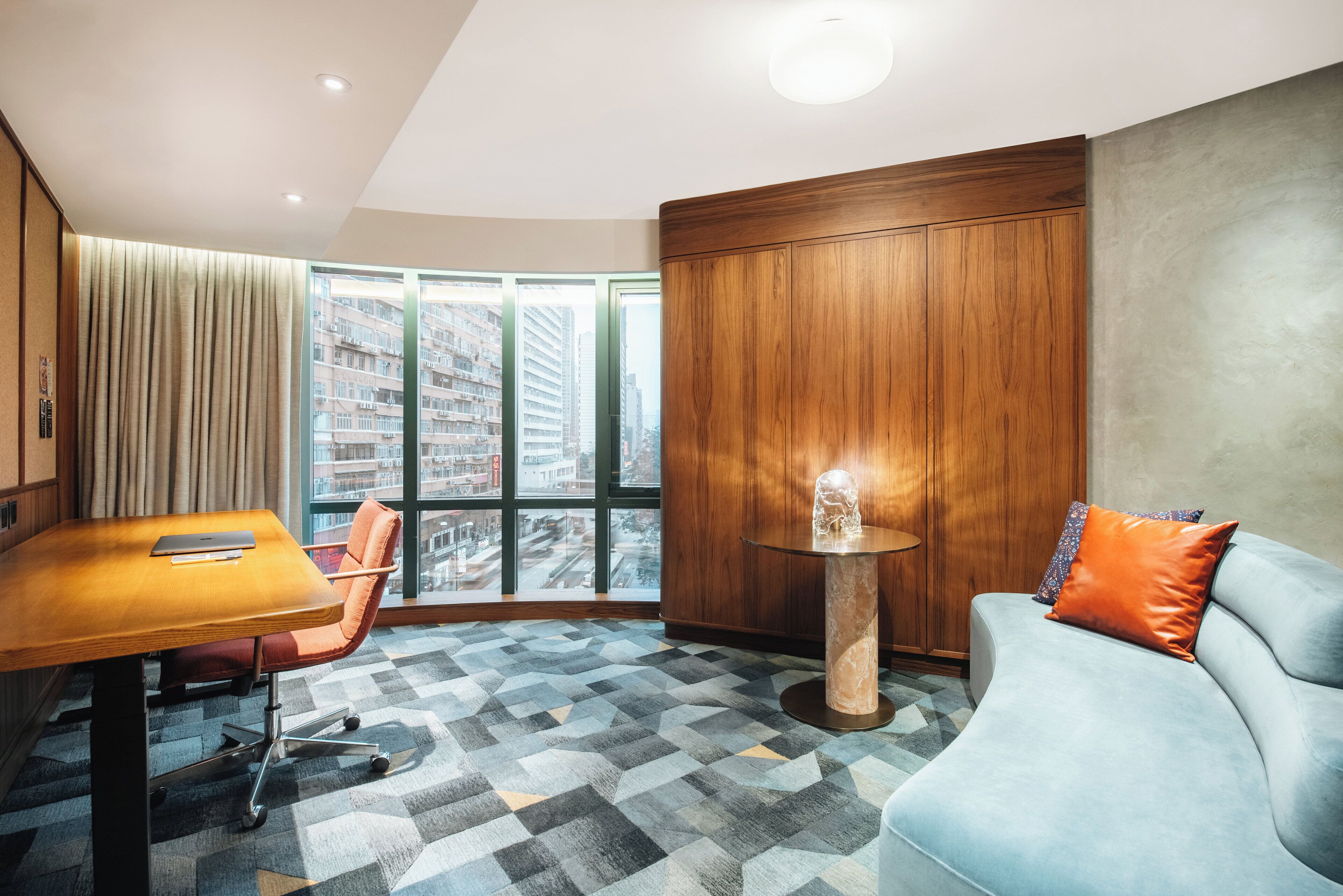 Une chambre d’hôtel moderne avec une grande fenêtre, une armoire en bois, une table ronde, un canapé bleu et un tapis à motifs.