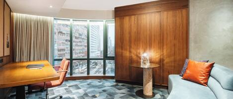 Une chambre d’hôtel moderne avec une grande fenêtre, une armoire en bois, une table ronde, un canapé bleu et un tapis à motifs.