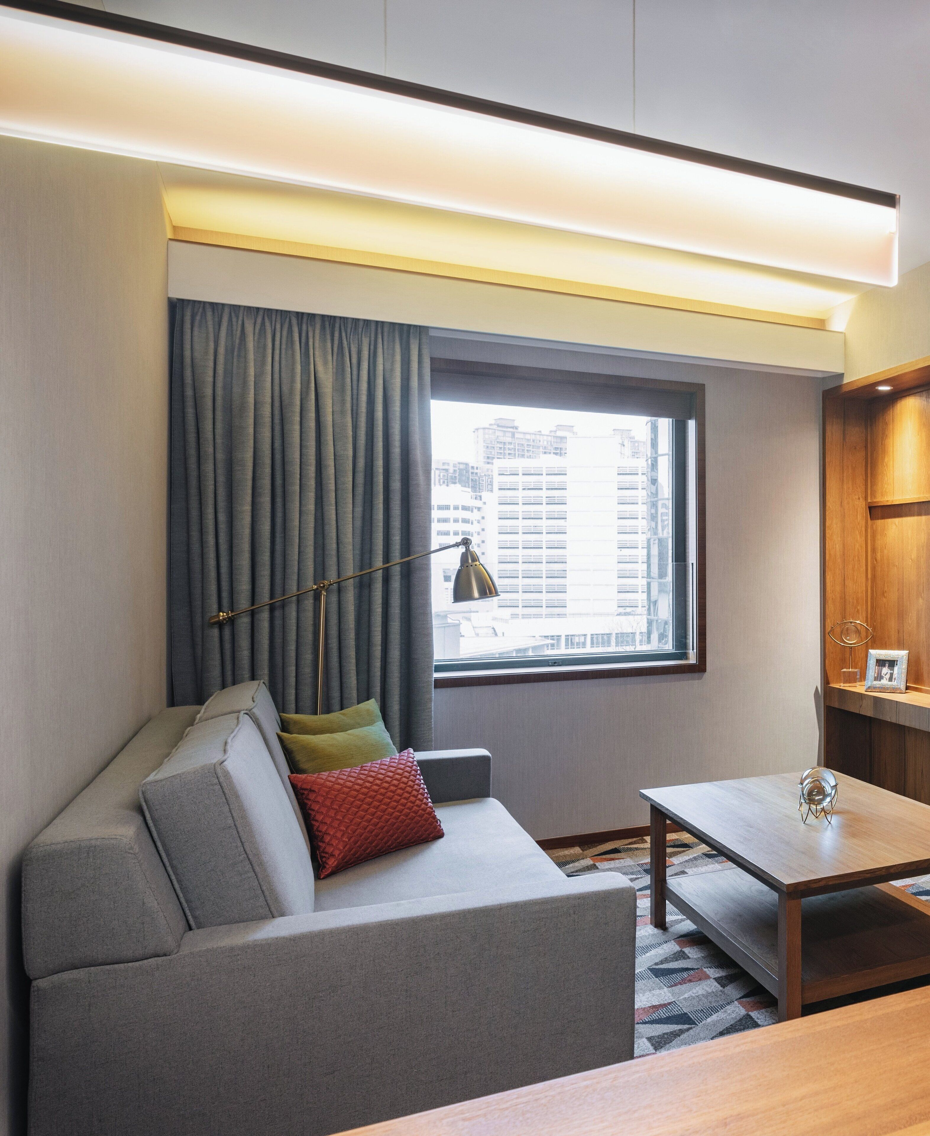 A modern hotel room with a grey sofa, a small table, a lamp, and a view of the city through a window with curtains.
