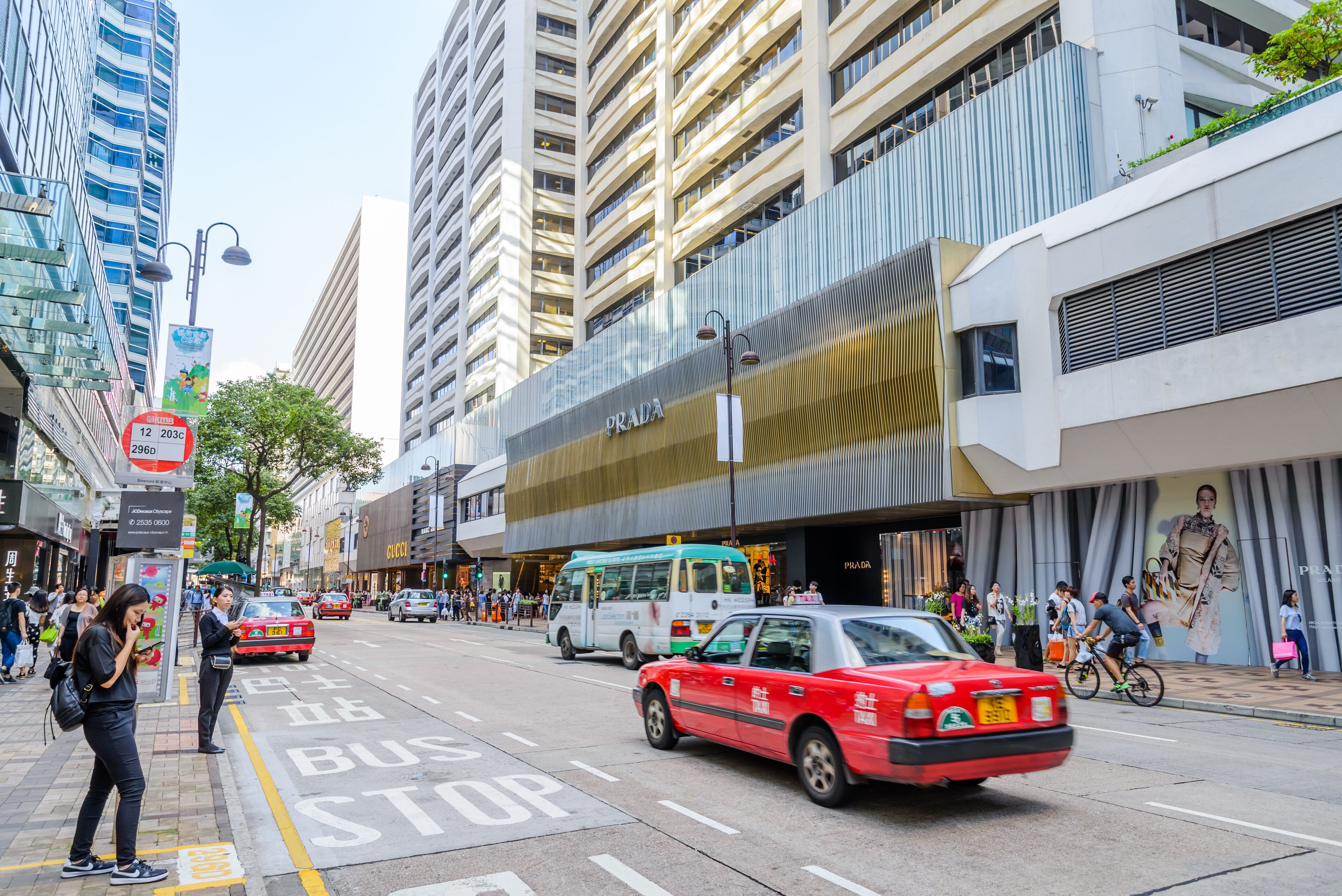 A busy urban street with a red taxi, a bus stop, and a Prada store.
