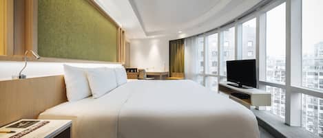 A modern hotel room with a large bed, a desk, a television, and a view of the city through large windows.