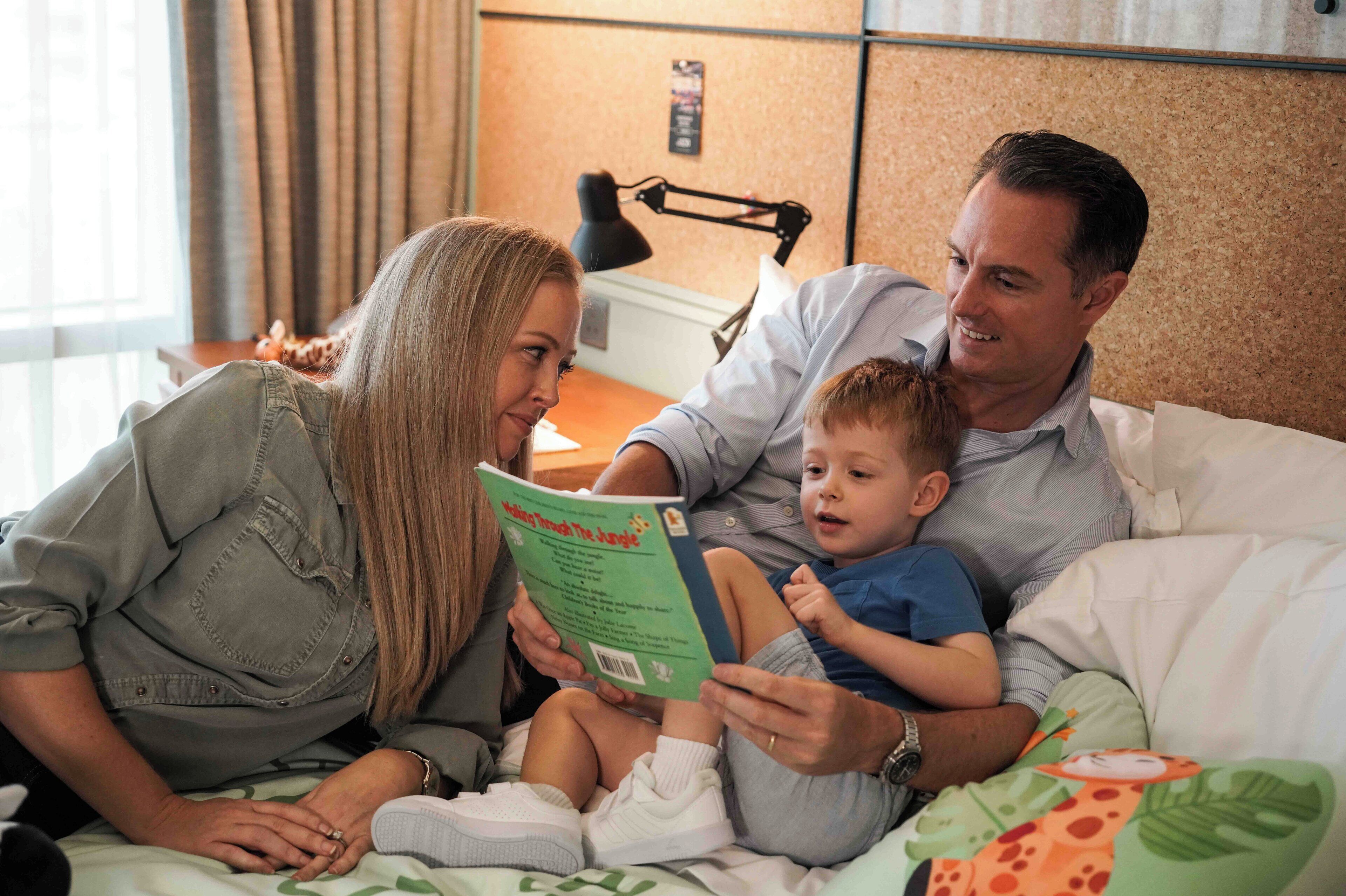 A family of four in a hotel room with a bed, a nightstand, and a lamp.