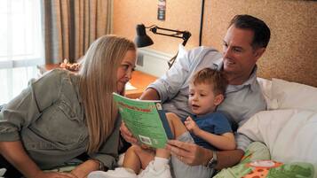 A family of four in a hotel room with a bed, a nightstand, and a lamp.