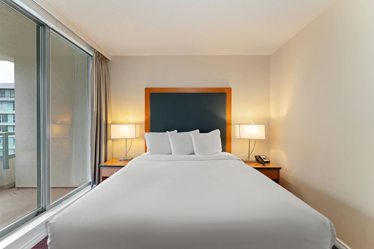 Une chambre d’hôtel avec un grand lit, deux tables de chevet avec des lampes, une fenêtre avec des rideaux et une vue sur un bâtiment à l’extérieur.
