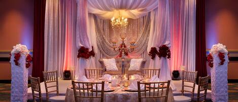 A banquet hall with a round table set for a wedding reception, surrounded by chairs, and decorated with floral arrangements and drapery.
