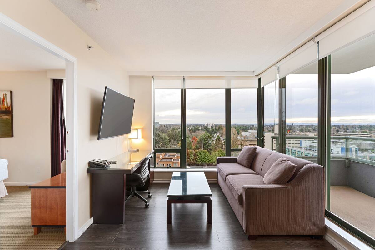Un salon moderne comprenant un canapé, une table basse, un bureau avec une chaise et un téléviseur fixé au mur. Une grande fenêtre offre une vue sur le paysage urbain.