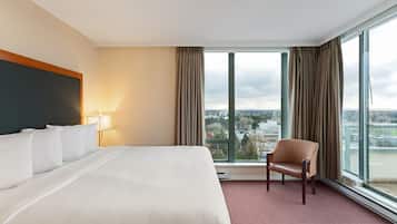A hotel room with a large bed, a chair, a lamp, and a view of a cityscape through large windows.