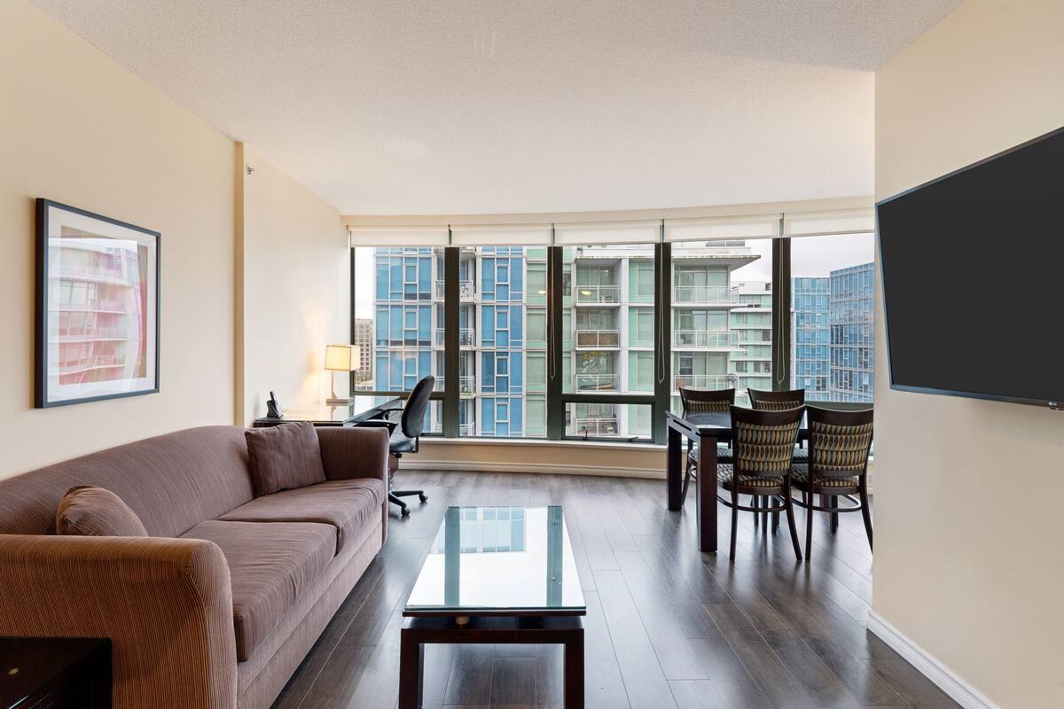 Un salon moderne comprenant un canapé, une table basse en verre, une table à manger avec des chaises et une grande fenêtre donnant sur la ville.