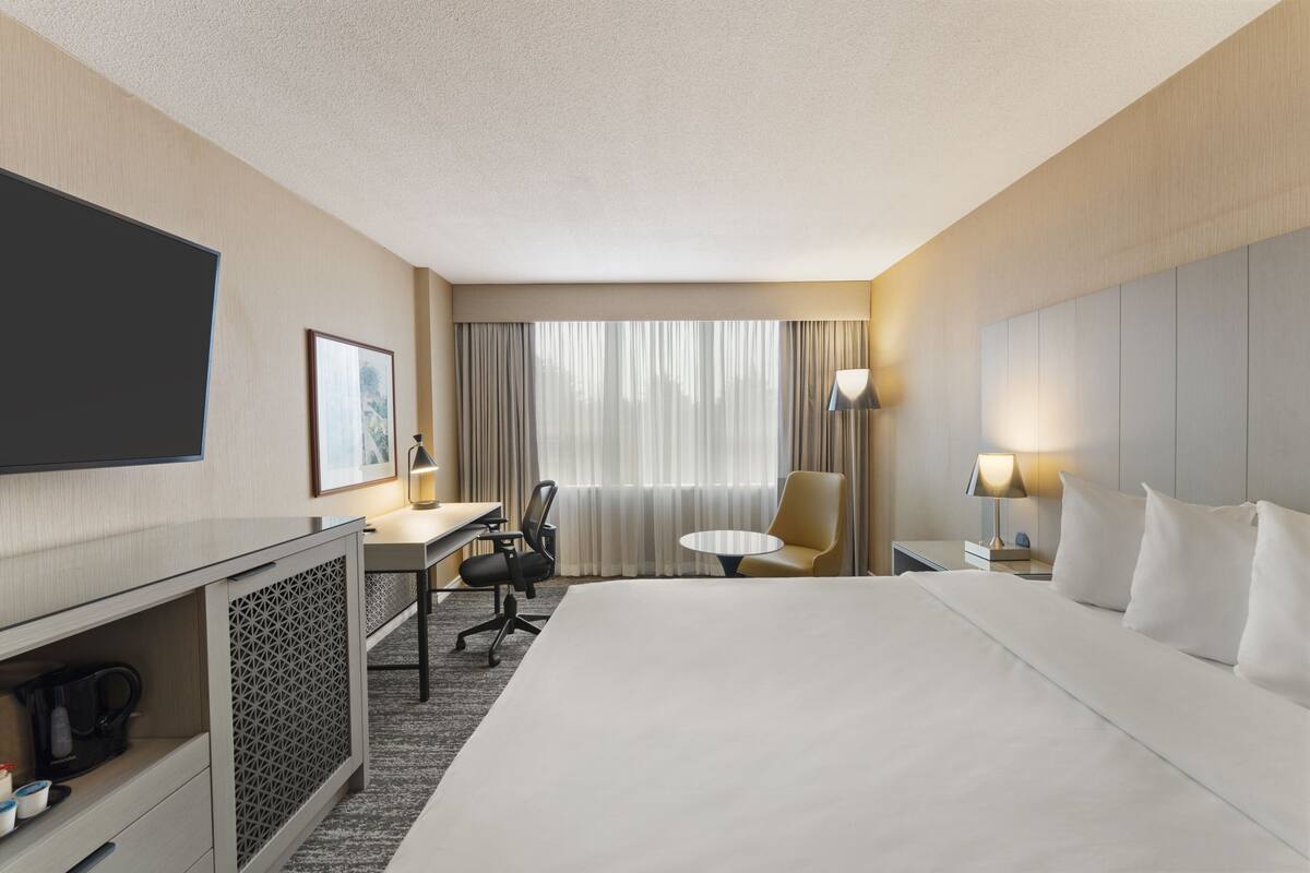 Une chambre d’hôtel avec un grand lit, un bureau avec une chaise, une télévision, une lampe et une fenêtre avec des rideaux.