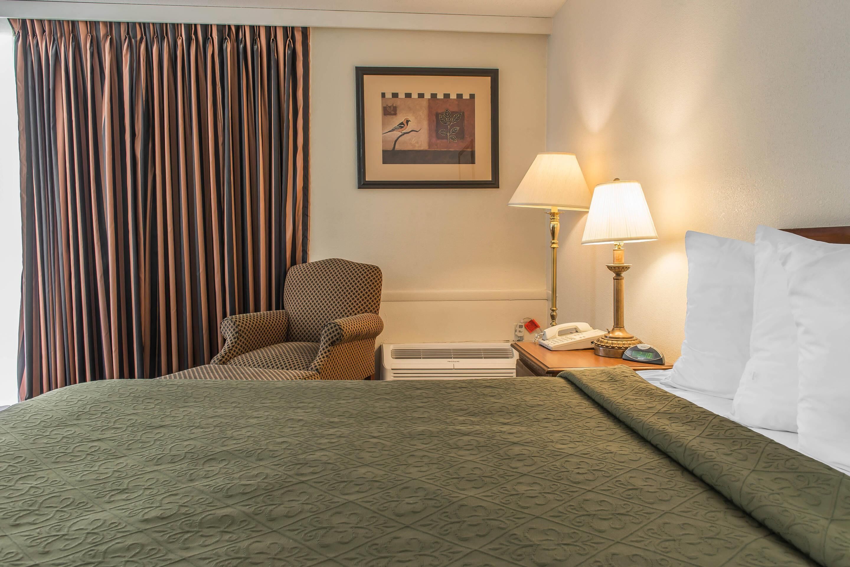 Habitación de hotel con cama, una silla, mesita de noche con lámpara, cuadro en la pared y ventana con cortinas.