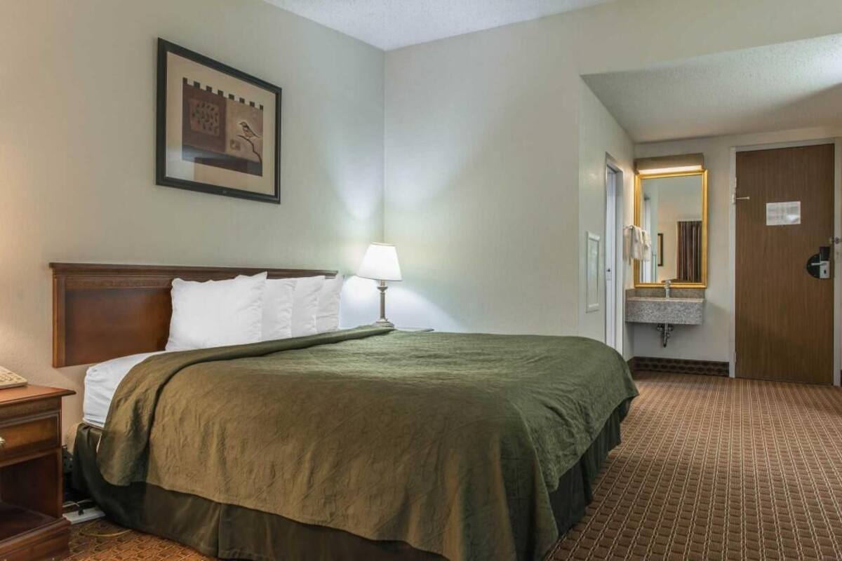 A hotel room with a bed, a nightstand, a lamp, a mirror, a sink, and a door.