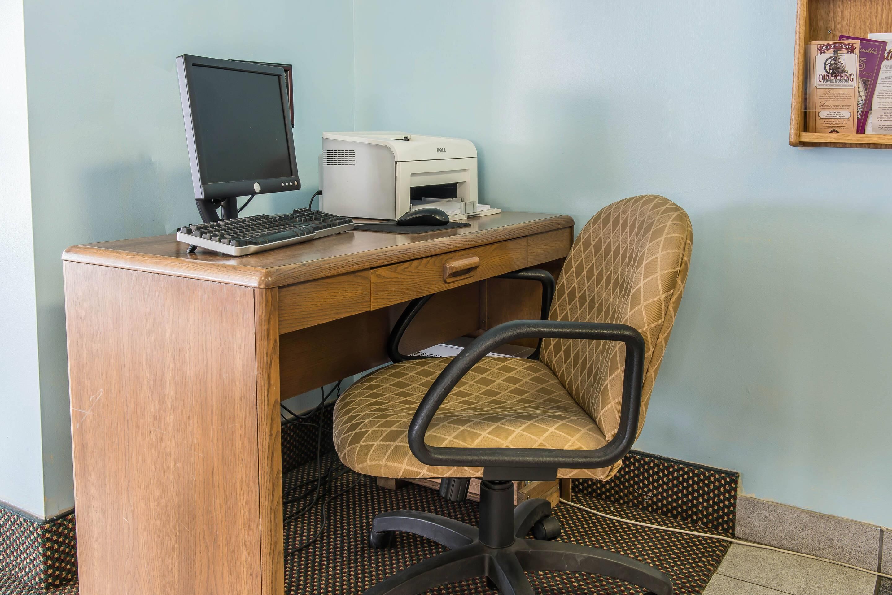 A wooden desk with a computer, keyboard, and monitor. There is a chair with a patterned cushion.