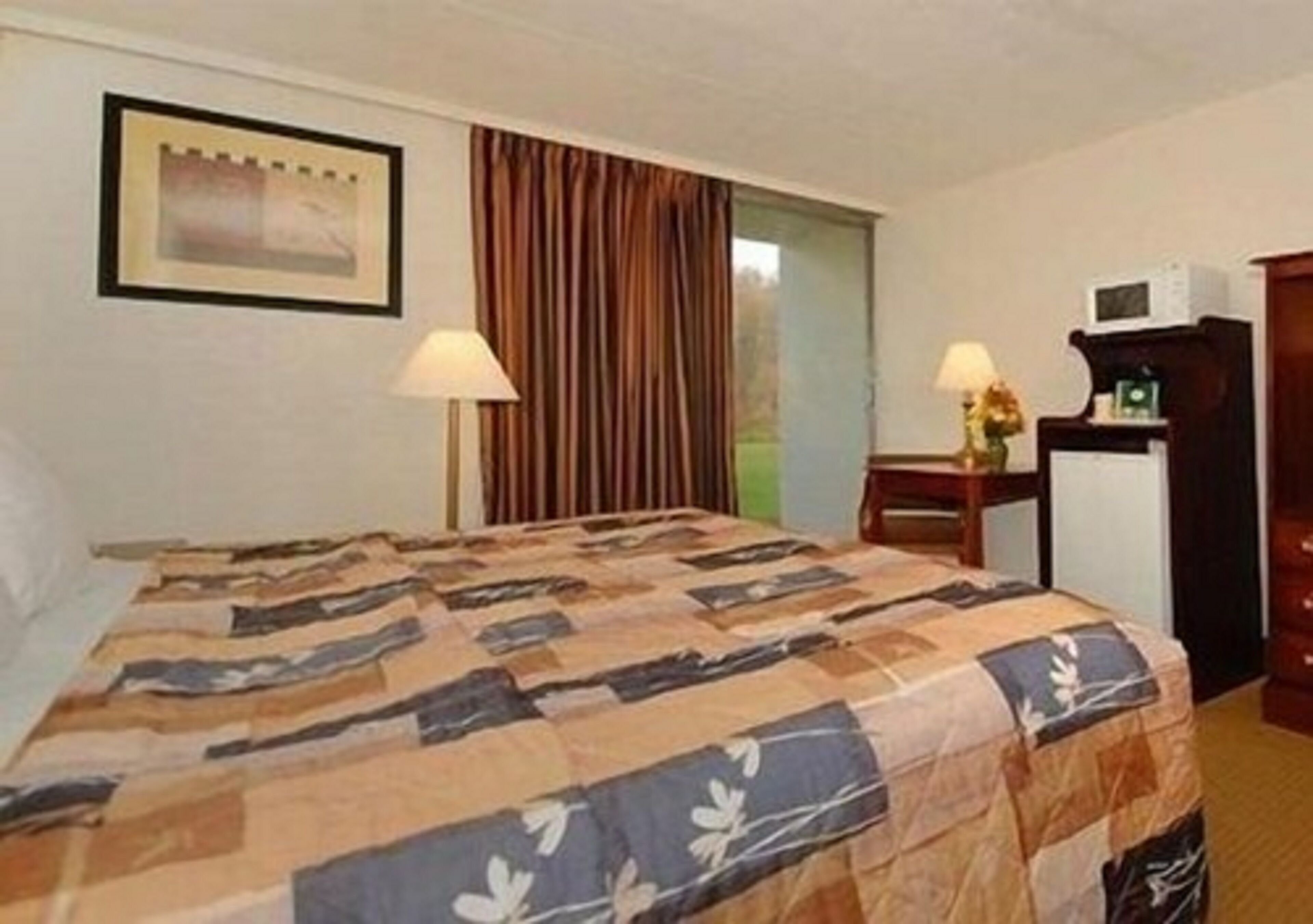 A hotel room with a bed, two bedside lamps, a desk with a microwave, and a framed picture on the wall.