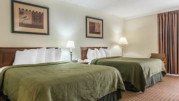 A hotel room with two beds, each with a green bedspread, white pillows, and a wooden headboard. There are two framed pictures on the wall, a lamp on each side of the beds, and a window with curtains.