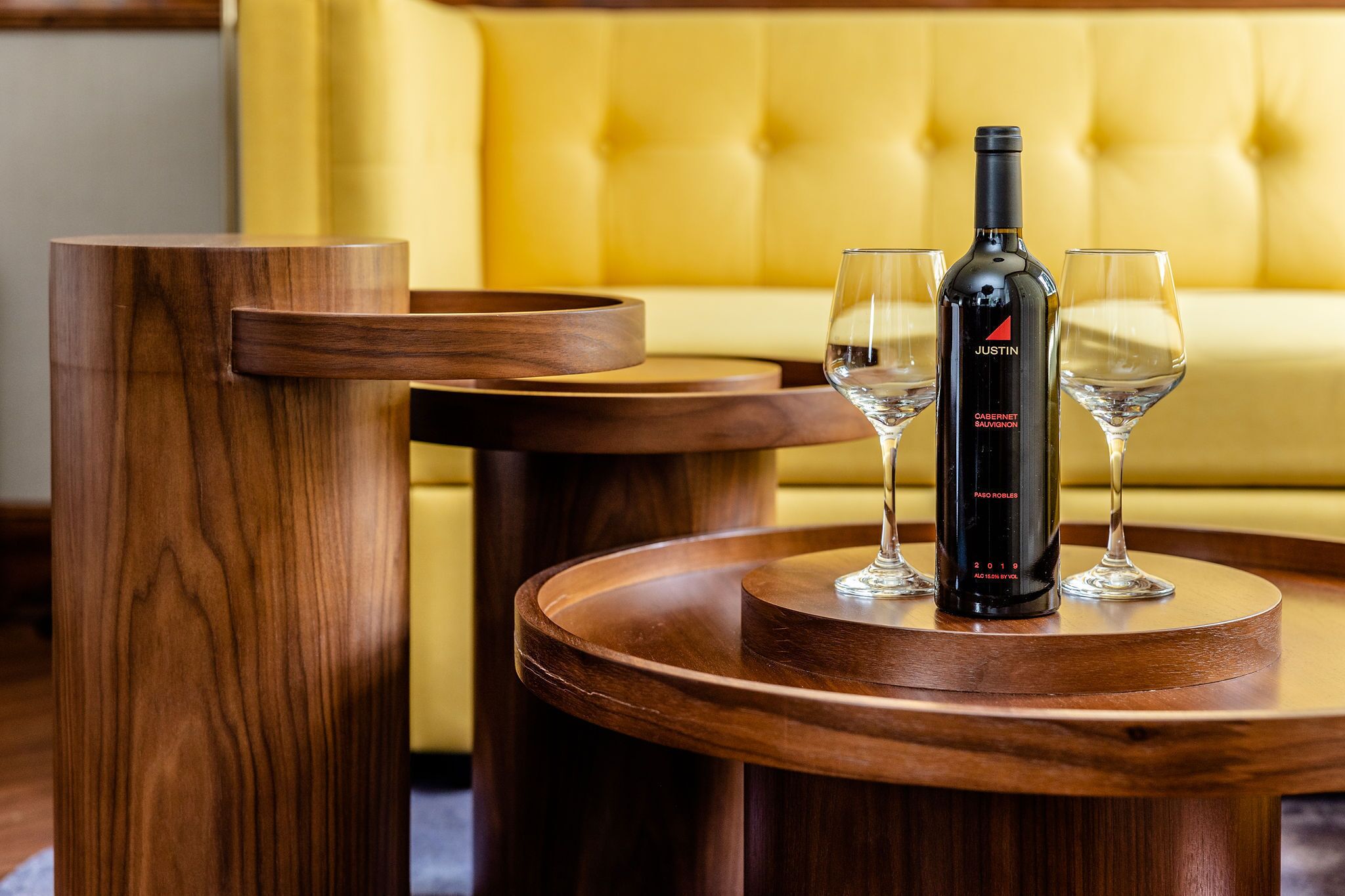 A wooden table with a bottle of wine and two glasses on it, set against a yellow upholstered sofa.
