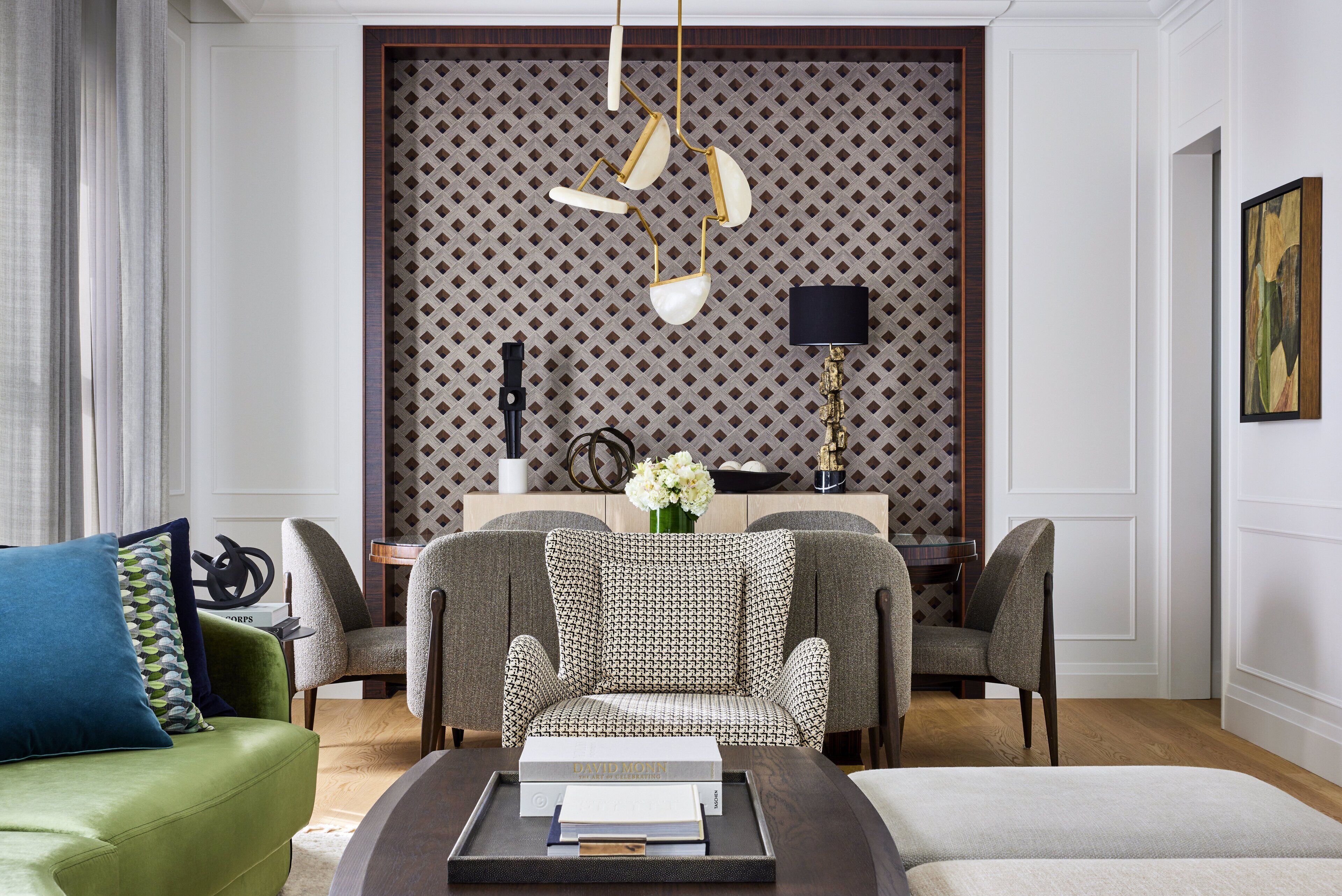 A modern living room with a patterned wallpaper, a green sofa, two armchairs, a coffee table with books, and a decorative lamp.