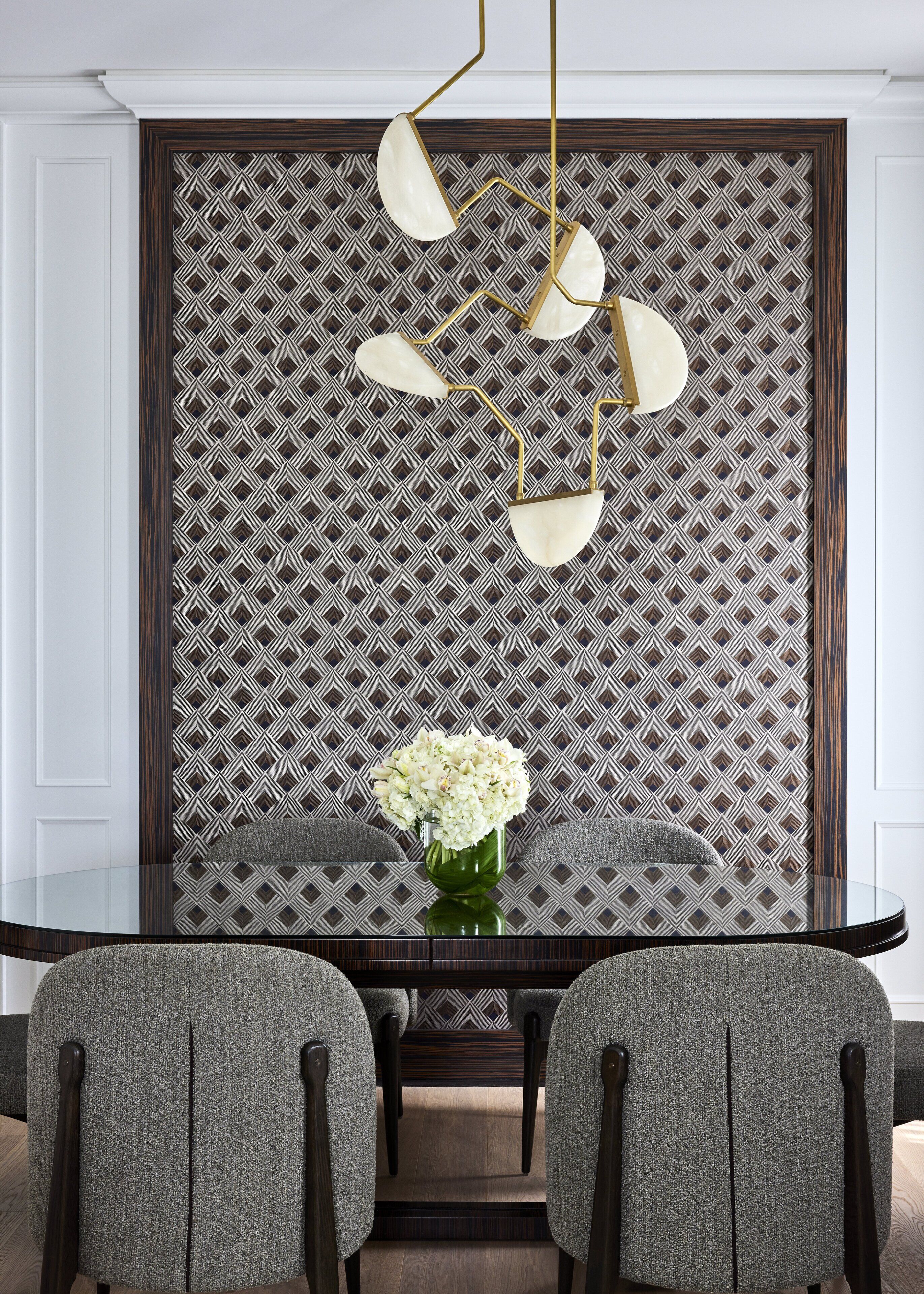 A dining area with a glass table, two grey upholstered chairs, a green vase with flowers, and a patterned wall with a geometric design.