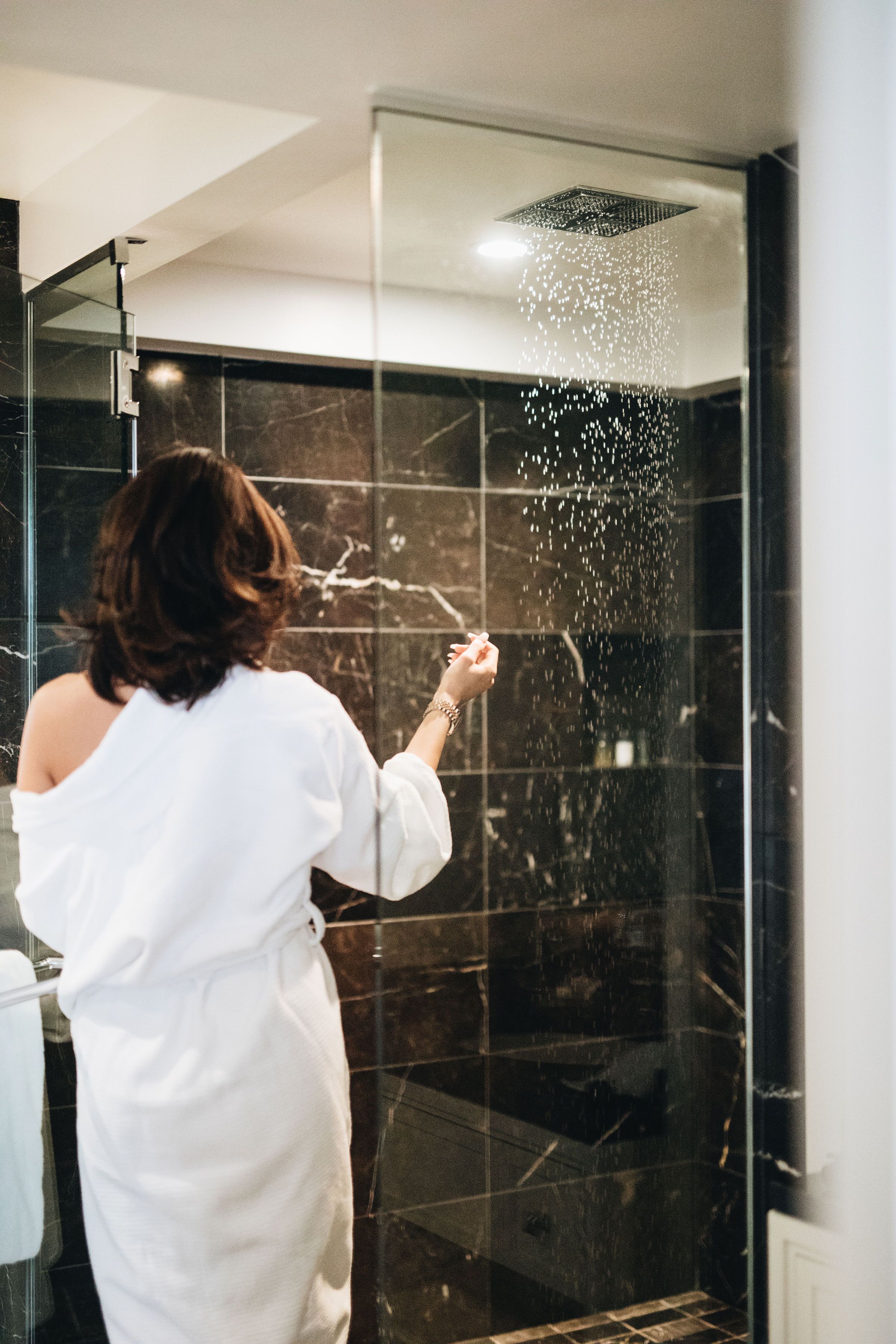 A woman in a white robe standing in a glass-enclosed shower area with a visible water spray.