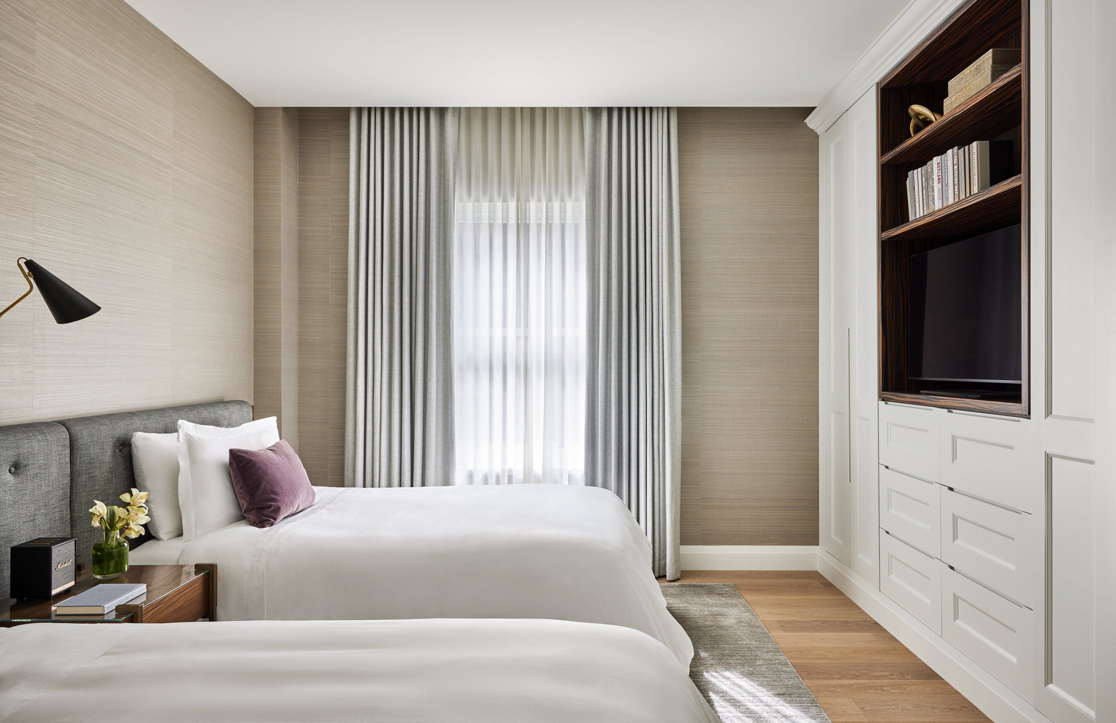 A modern hotel room with two beds, a large window with sheer curtains, a flat-screen TV, and a built-in bookshelf.
