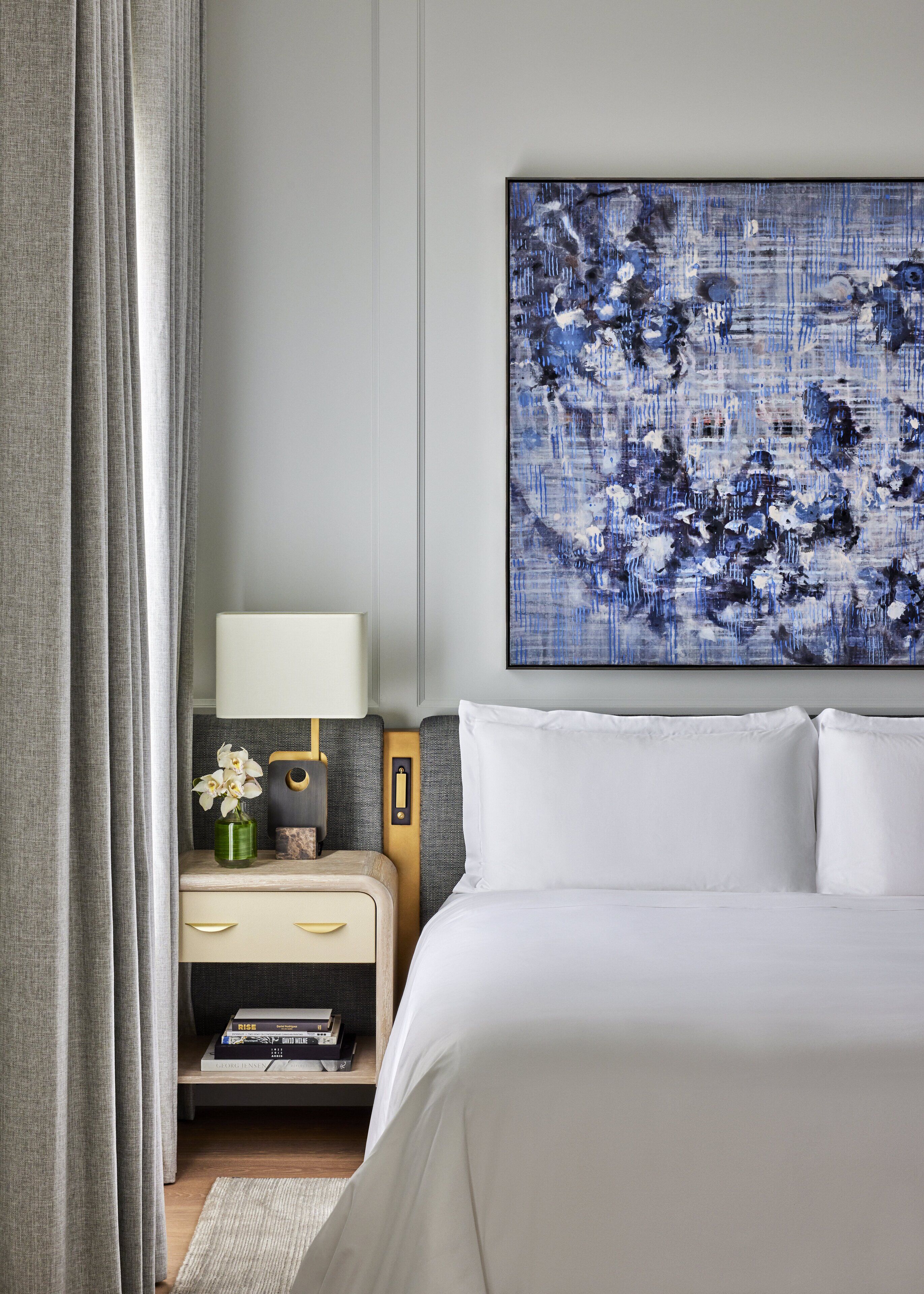A hotel room with a bed, a nightstand, a lamp, a painting, and a window with curtains.