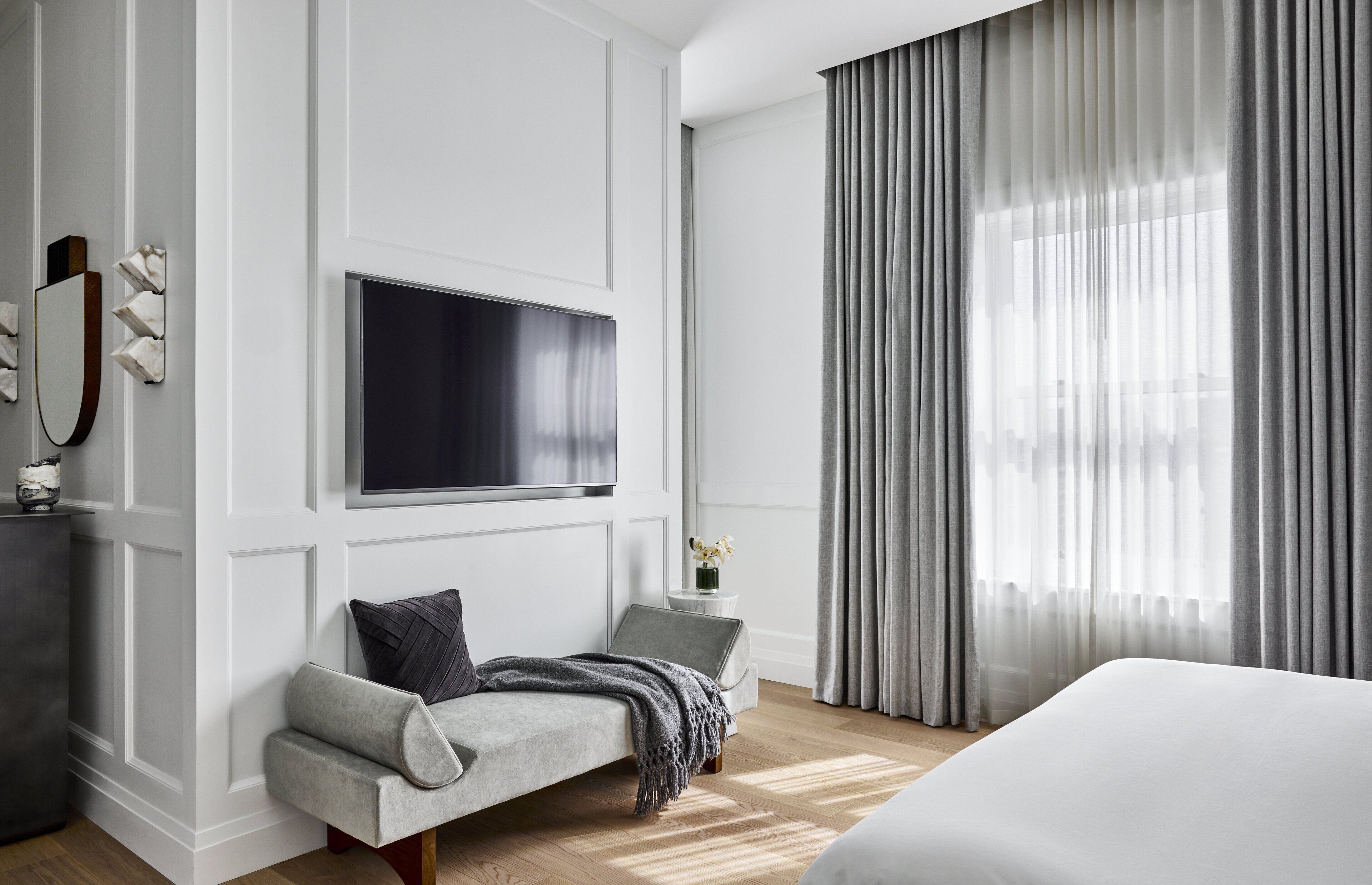 Een moderne hotelkamer met een groot bed, een flatscreen televisie aan de muur, een zithoek met een bank en een groot raam met dunne gordijnen.
