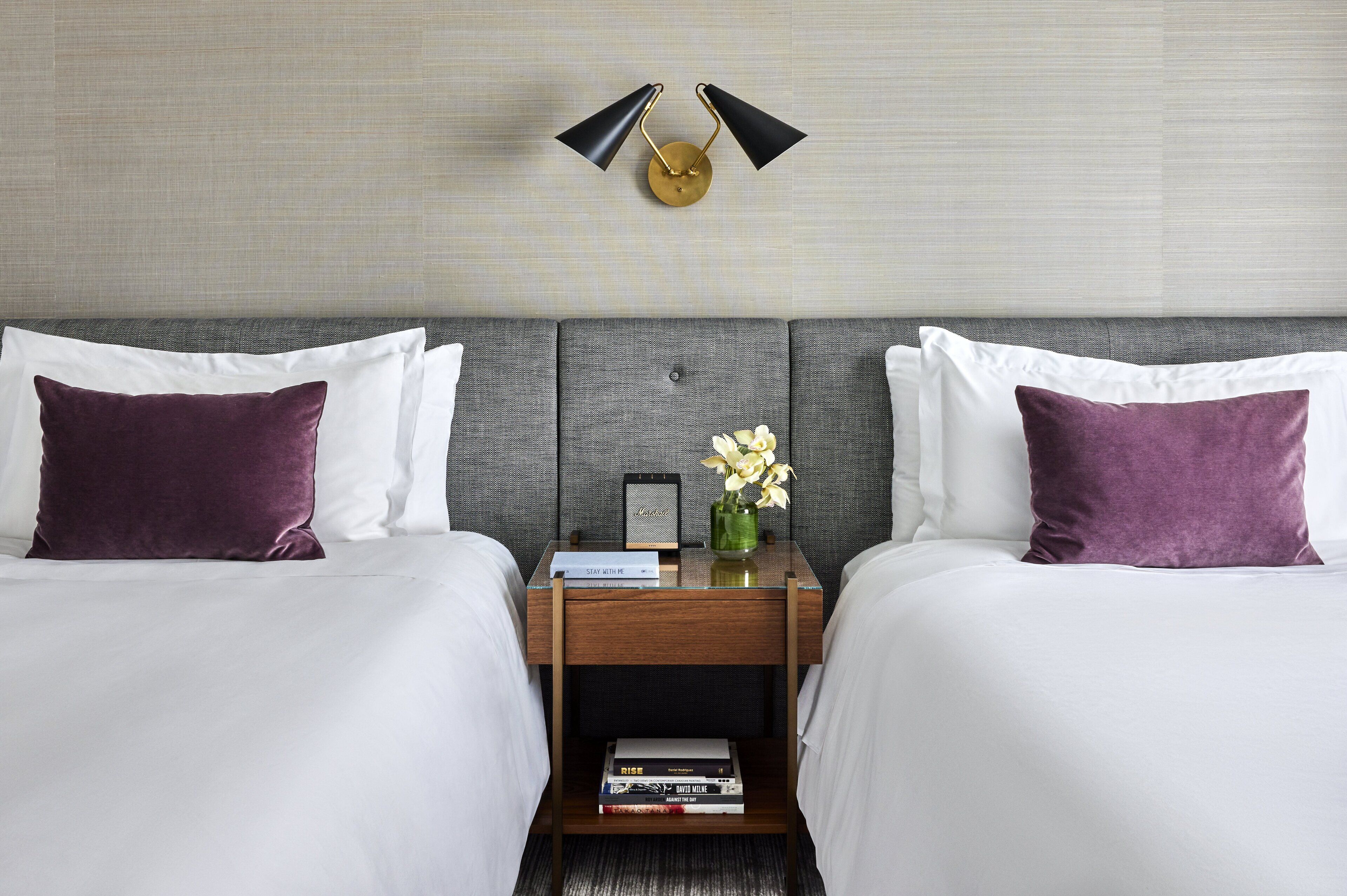 Habitación de hotel con dos camas, una mesita de noche con lámpara, un jarrón con flores y una pequeña mesa con un libro y un reloj.
