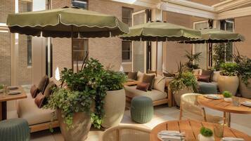 A well-lit outdoor seating area with multiple tables and chairs, surrounded by large potted plants and umbrellas.