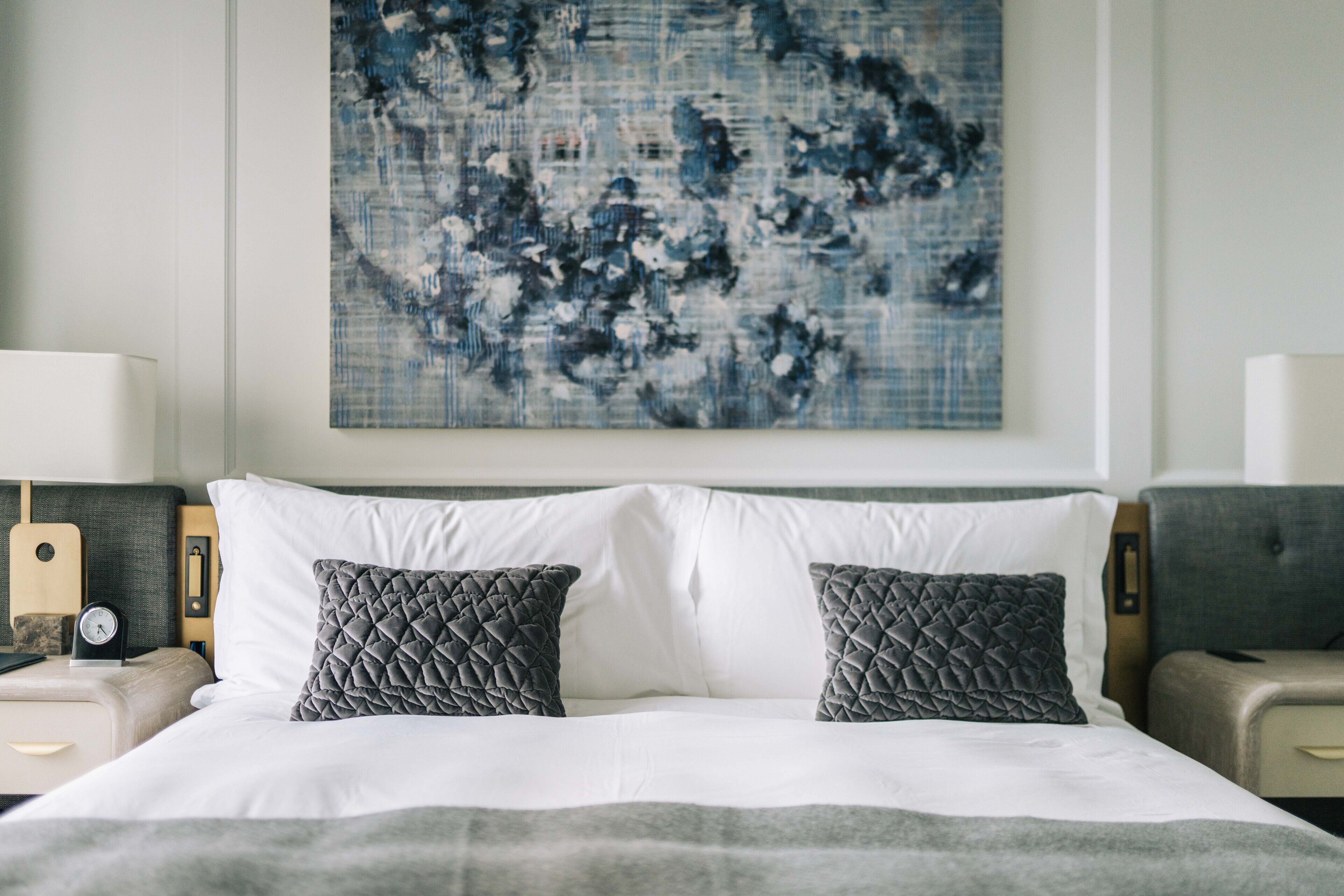 Un lit bien fait, avec du linge de lit blanc et deux oreillers gris, une tête de lit en bois, une table de chevet avec une lampe et un tableau abstrait accroché au mur.