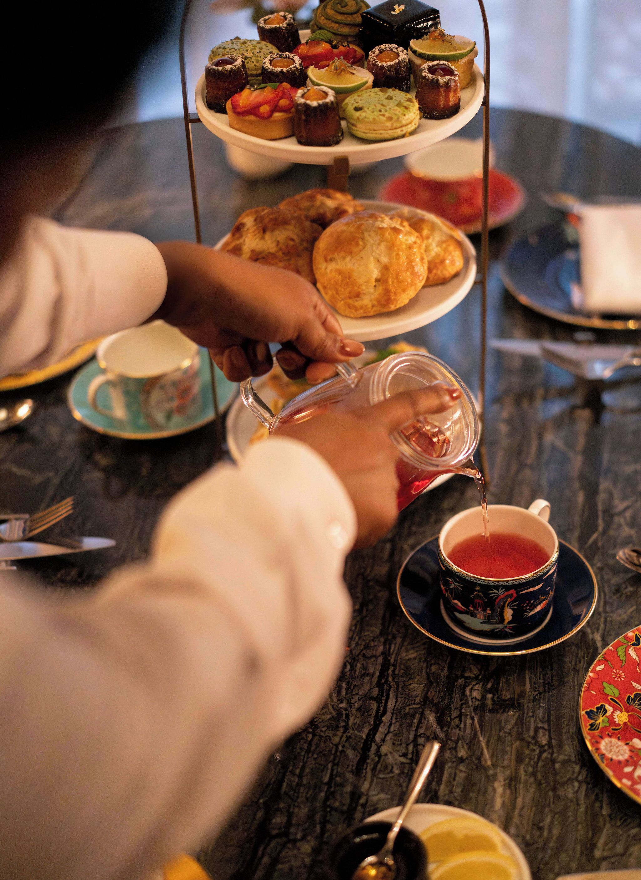 Una persona sirviendo té en una taza, un soporte de varios niveles para postres y diferentes platos y utensilios sobre una mesa.