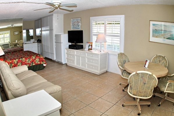 A compact room with a bed, a desk, a television, a dining table with chairs, and a ceiling fan.