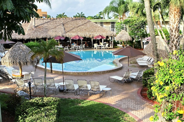 A pool area with lounge chairs and umbrellas, surrounded by palm trees and a thatched-roof building.