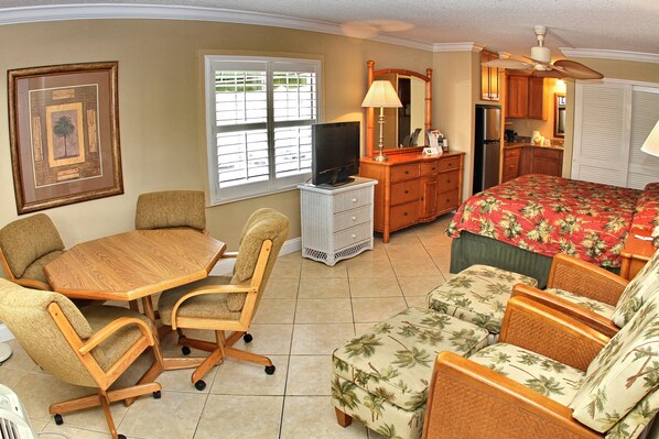 A compact hotel room with a dining table, two chairs, a sofa, a TV, a dresser, a mirror, and a kitchenette.