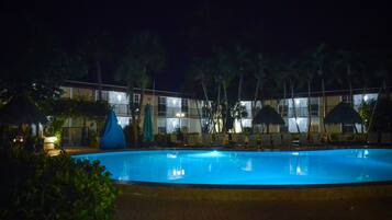 A swimming pool with lounge chairs and umbrellas, surrounded by palm trees and a building with multiple balconies.