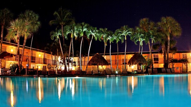 A nighttime view of a hotel pool area with palm trees and thatched-roof structures. - Gulfcoast Inn Naples (Naples)