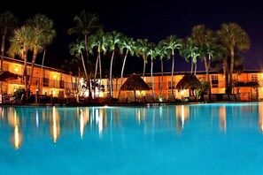 A nighttime view of a hotel pool area with palm trees and thatched-roof structures.