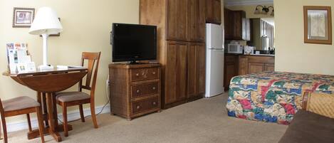 A hotel room with a dining table, chairs, a television, a wardrobe, a refrigerator, a bed with a colorful quilt, and a kitchen area.
